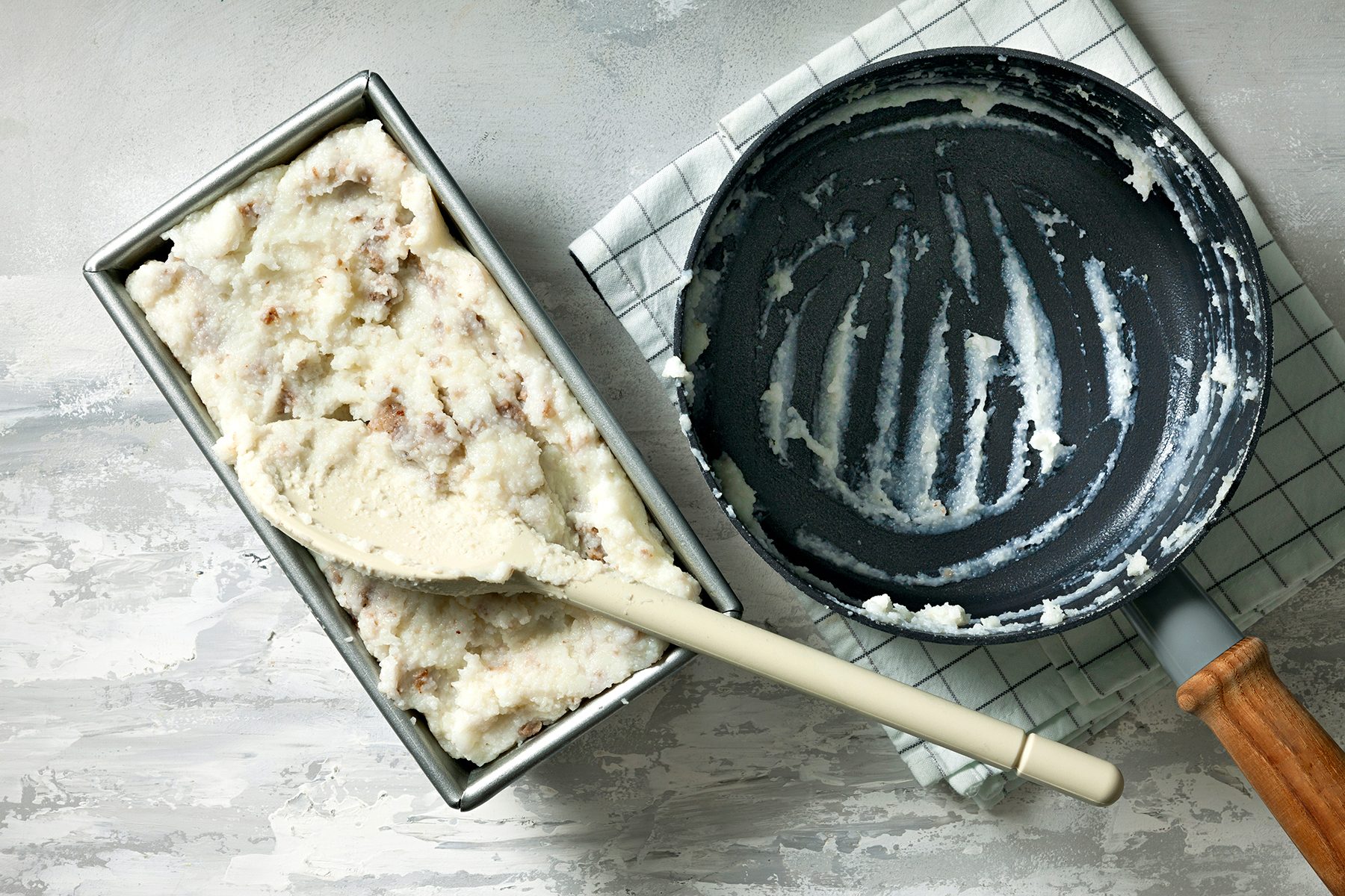 A rectangular metal container filled with mashed potatoes sits next to an empty, used frying pan with remnants of mashed potatoes. A white spatula rests on the container. Both are on a checkered cloth on a marbled surface.