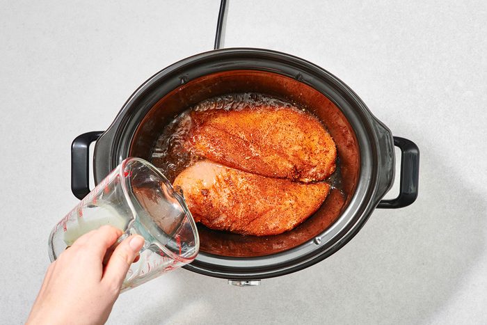 Pouring chicken broth into the slow cooker with the seasoned chicken breast