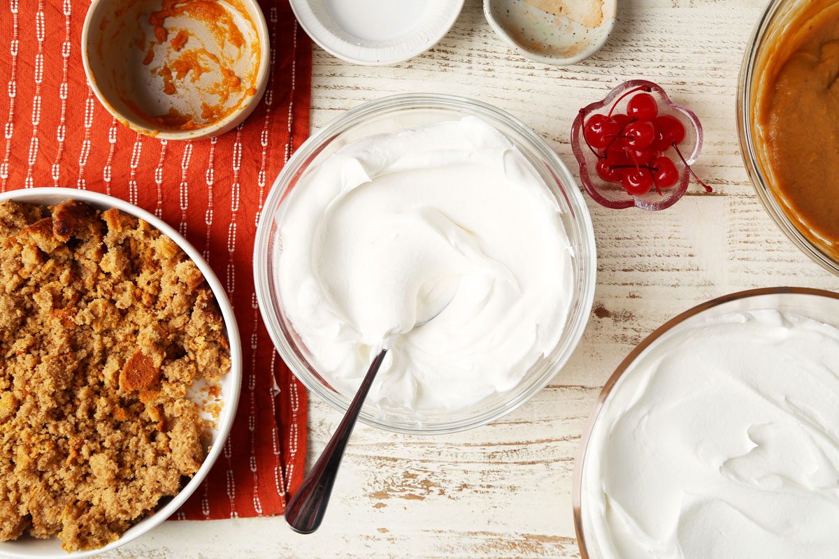Process of making Taste of Home's Pumpkin Trifle layered in a glass serving bowl on a white wooden surface.
