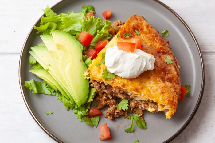 Close up of single serving of Taste of Home Taco Pie on a white wood surface, lettuce, tomato, avocado, cilantro, sour cream