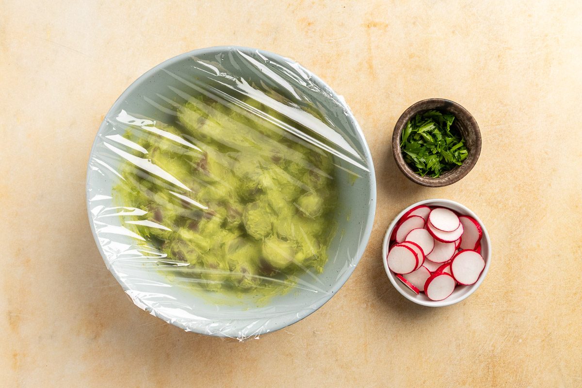 Taste of Home Vegan Potato Salad recipe photo of the salad in a mixing bowl covered with plastic wrap to be set in the fridge. Radishes and parlsely are on the side.