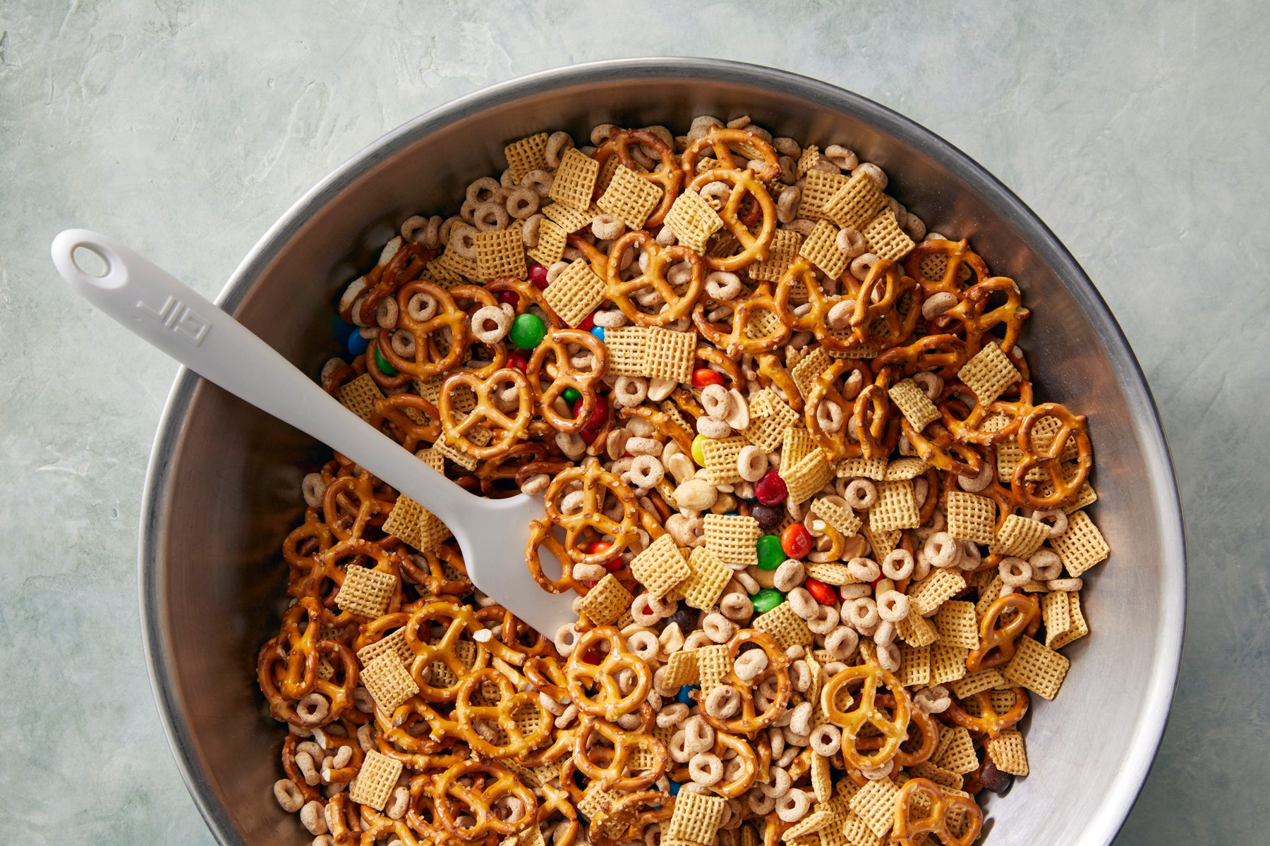 overhead shot; white background; A large bowl contains a mixture of various snack foods, including pretzels, Chex cereal, and colorful candy; A spatula rests in the bowl, suggesting that the ingredients are being combined