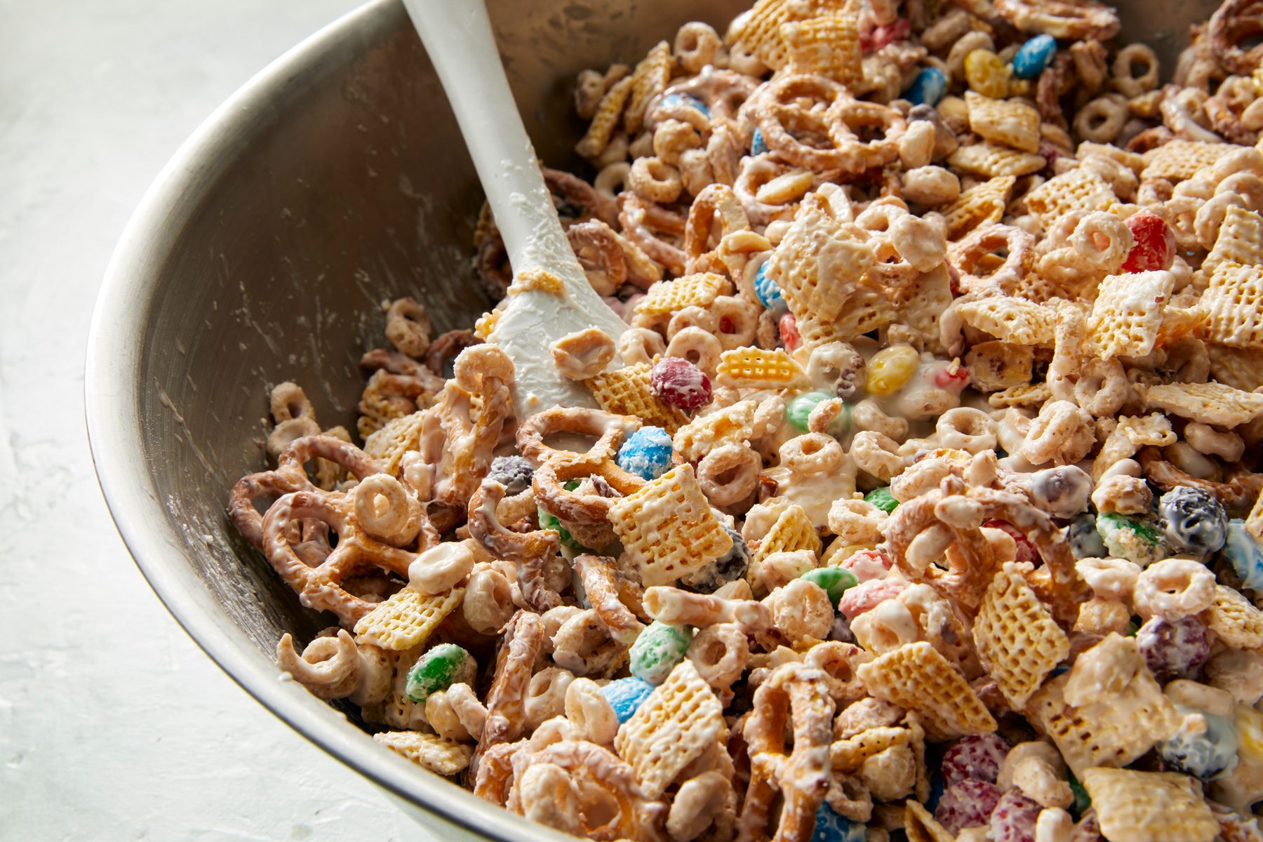 3/4th shot; close shot; white background; A large bowl contains a mixture of various snack foods, including pretzels, Chex cereal, and colorful candy; A spatula rests in the bowl, suggesting that the ingredients are being combined