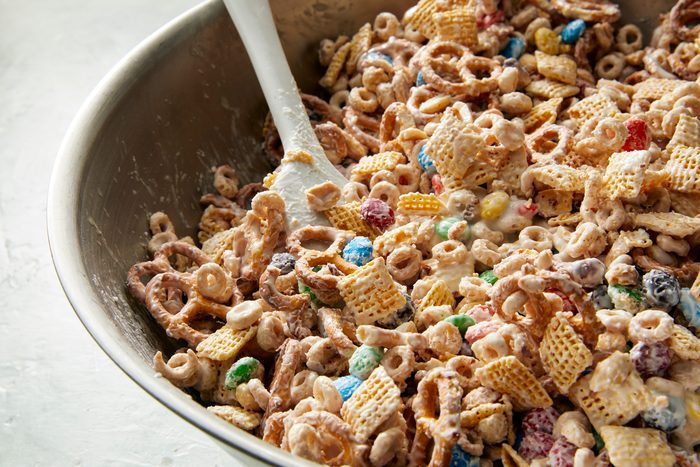 3/4th shot; close shot; white background; A large bowl contains a mixture of various snack foods, including pretzels, Chex cereal, and colorful candy; A spatula rests in the bowl, suggesting that the ingredients are being combined