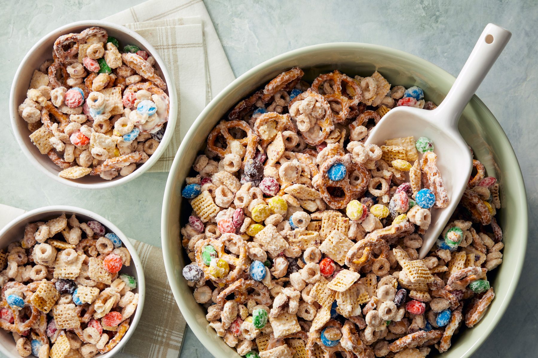 overhead shot; white background; A large bowl filled with a colorful mixture of pretzels, Chex cereal, and candy is the focal point of the image; A white scoop rests in the bowl, ready for serving; Smaller bowls filled with the same mixture are arranged around the larger bowl, placed over kitchen towel
