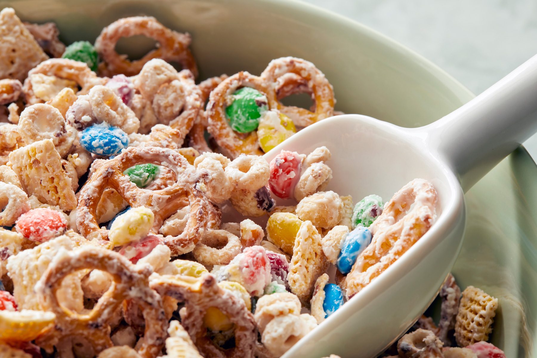 close shot; white background; A large bowl filled with a colorful mixture of pretzels, Chex cereal, and candy is the focal point of the image; A white scoop rests in the bowl