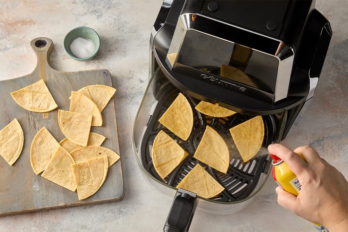 overhead shot of an air fryer with a basket full of tortilla chips, A person is spraying the chips with cooking spray, There is a wooden cutting board to the left with more tortilla chips;