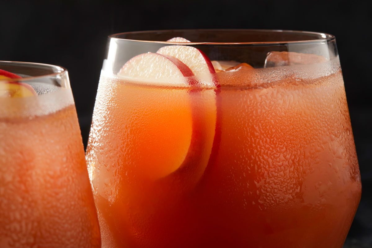 a close-up of a refreshing Bourbon Apple Cider Cocktail, featuring a vibrant orange-red hue and a chilled glass, The drink is garnished with slices of apple