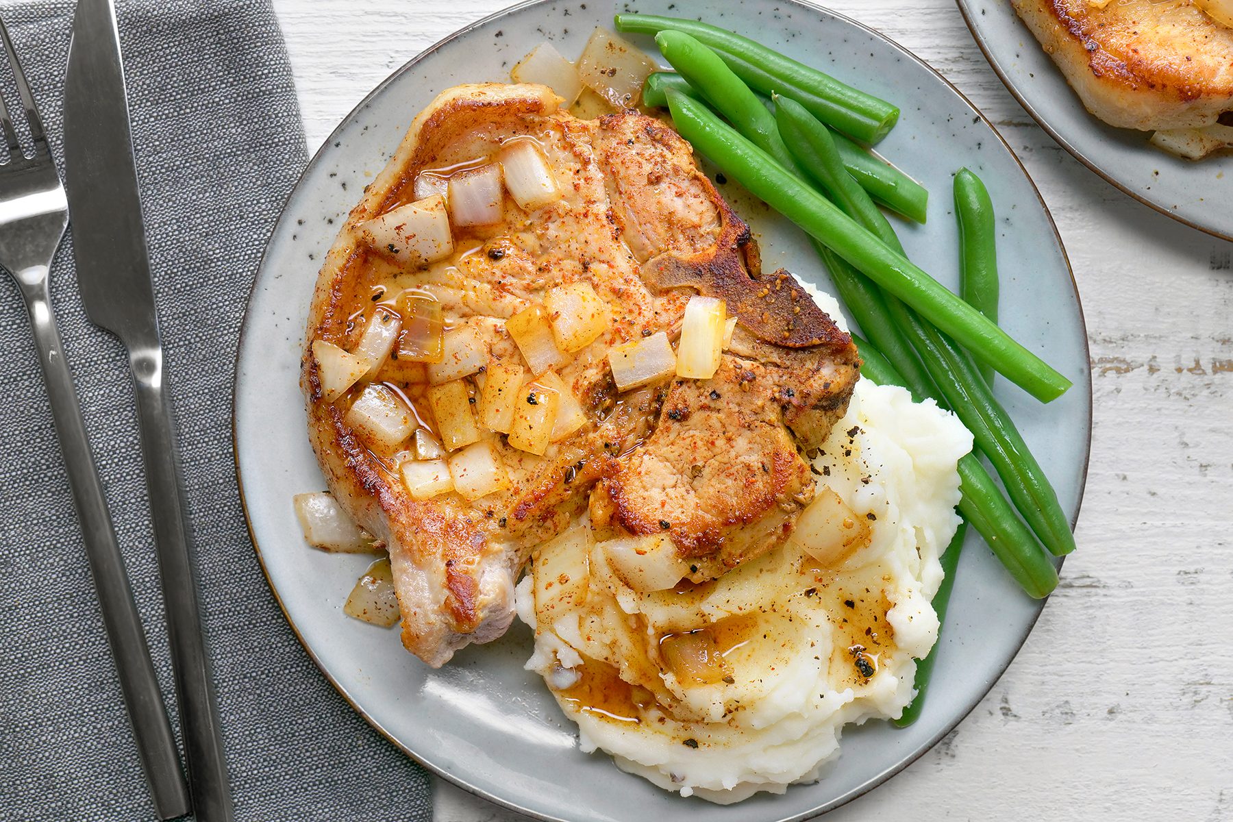 A plate with a seasoned pork chop topped with diced onions, served alongside a portion of creamy mashed potatoes and steamed green beans. A fork and knife are placed next to the plate on a gray napkin.