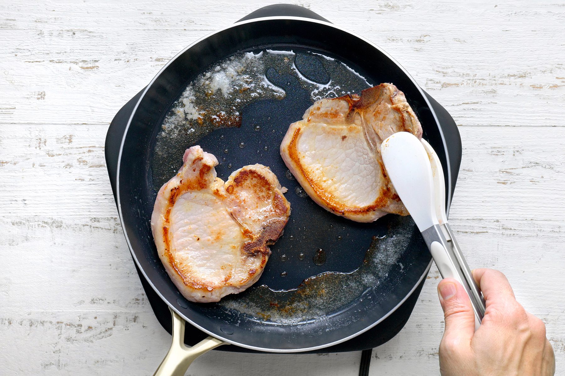 A hand uses tongs to flip one of two pork chops cooking in a frying pan. The meat is sizzling, with visible browning, on a white wooden surface background.