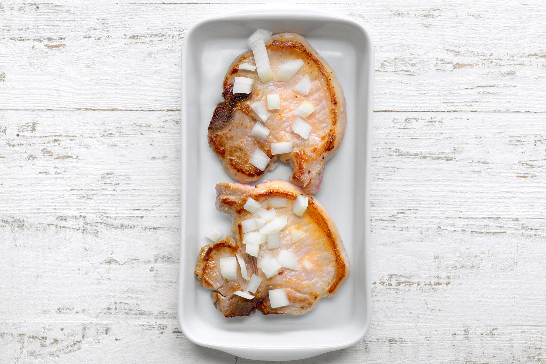Two cooked pork chops topped with chopped onions are placed on a white rectangular dish against a white wooden background.