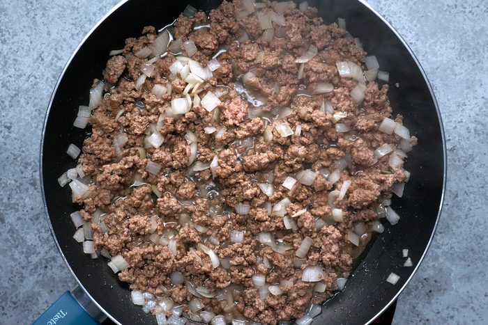 Ground beef and chopped onions sizzling in a black frying pan. The mixture is lightly browned, with small bubbles indicating it’s cooking on a stovetop. The background features a gray countertop.