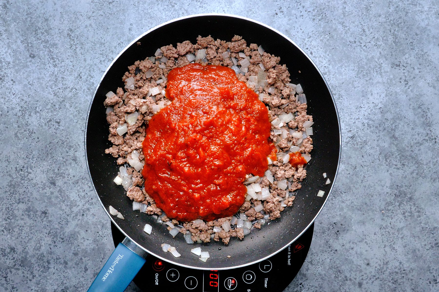 A frying pan on a countertop contains browned ground beef, chopped onions, and tomato sauce. The ingredients are ready to be mixed together, with the sauce positioned in the center.