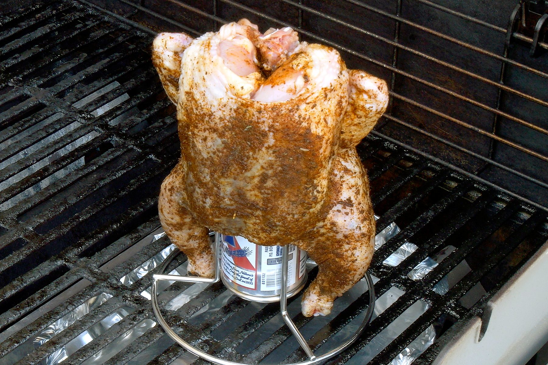 A seasoned whole chicken is being cooked upright on a grill, with a can placed inside it. The grill has metal grates and some aluminum foil underneath the chicken.