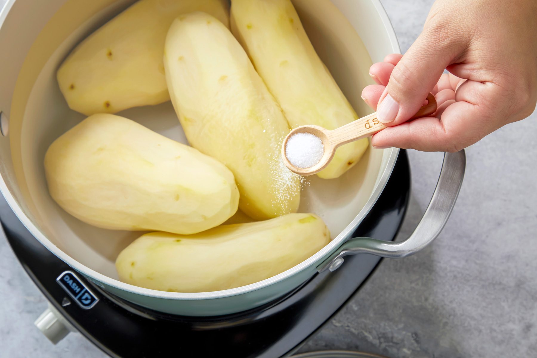 A stock pot filled with whole potatoes submerged in water, with a measuring spoon showing 1 teaspoon of salt beside it. The water is visibly covering the potatoes.