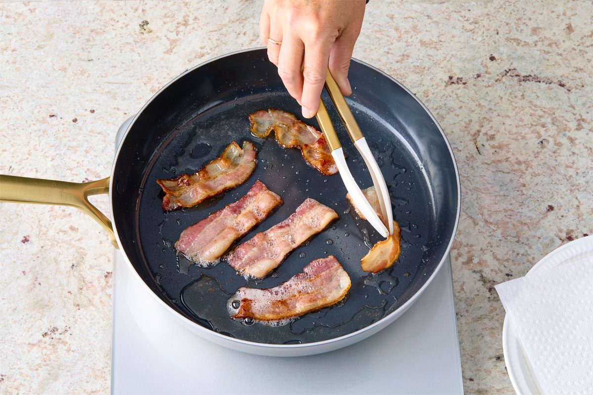 High angle view shot of a large skillet; cook bacon over medium heat until crispy; 7-8 minutes; stirring frequently; use a slotted spoon to transfer bacon to a paper towel-lined plate; discard all but 1 tablespoon bacon fat; tongs; marble surface;