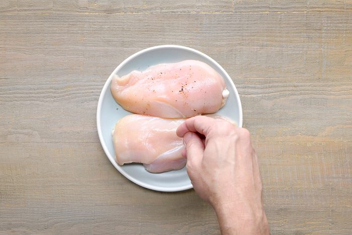overhead shot of two raw chicken breasts placed on a white plate, The chicken breasts are a pale white color and have a slightly moist texture. A person's hand is touching one of the chicken breasts, The chicken breasts are seasoned with a sprinkle of black pepper;