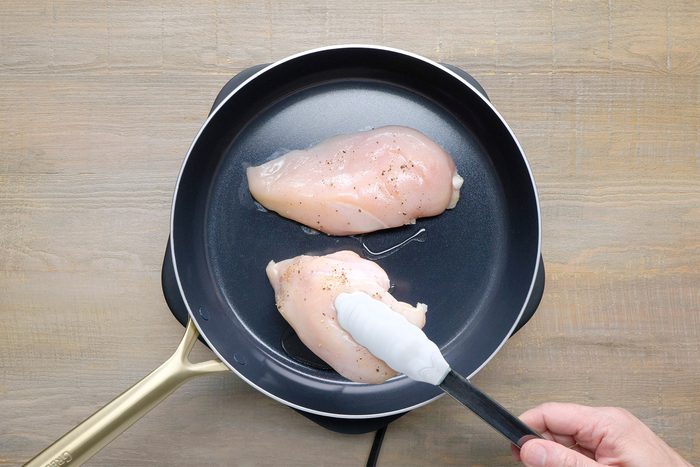 overhead shot of two chicken breasts cooking in a black skillet, The chicken breasts are a pale white color and are seasoned with black pepper, A spatula is being used to flip one of the chicken breasts;