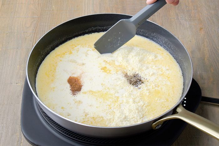 3/4th shot of a black skillet on an electric induction cooktop contains a creamy, pale yellow sauce, A person's hand is using a spatula to stir a mixture of spices into the sauce, The spices are a variety of colors, including brown and white, The sauce is simmering gently, and steam is rising from the skillet;