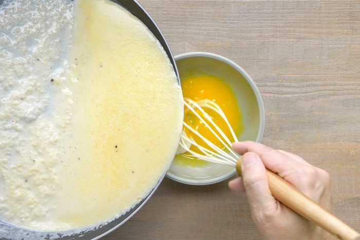 overhead shot of a person whisking an egg yolk in a small bowl, A skillet filled with a creamy, pale yellow sauce is visible in the background;