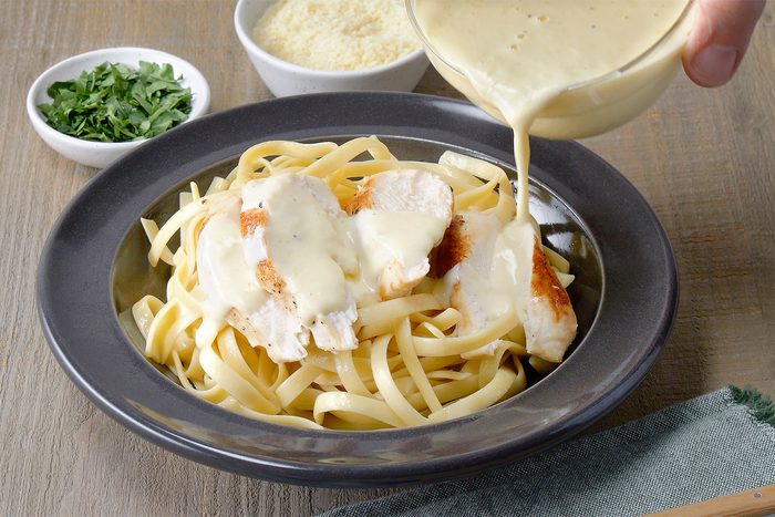 3/4th shot of a plate of Chicken Alfredo, Fettuccine pasta is coated in a creamy, pale yellow Alfredo sauce, with pieces of grilled chicken on top; The chicken is seasoned with herbs and has a golden brown color;