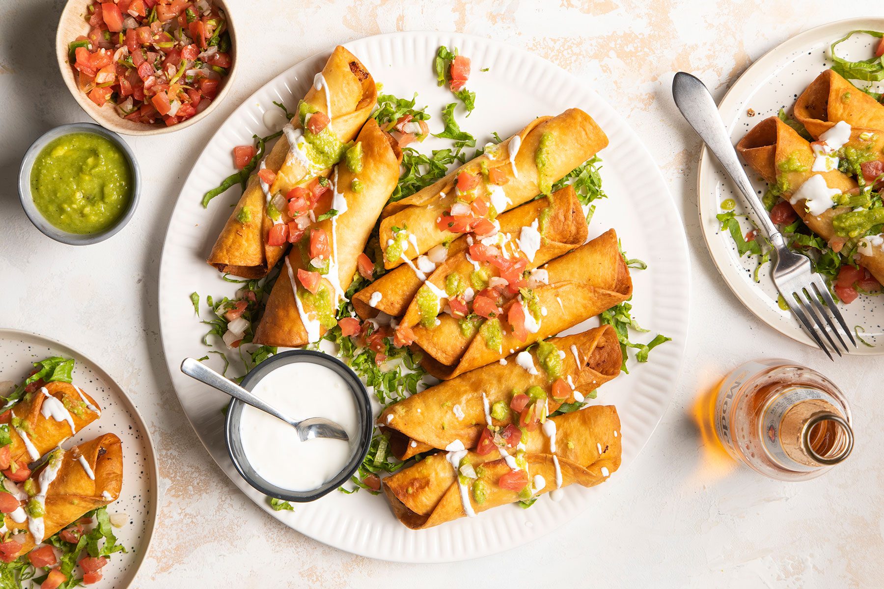 A plate of crispy taquitos topped with diced tomatoes, shredded lettuce, and drizzled with sauce. Small bowls of guacamole and sour cream are on the side. Another plate of taquitos and a drink are partially visible.