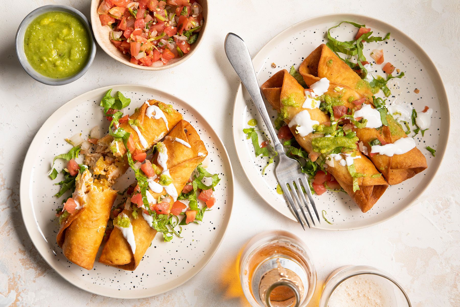 Two plates of crispy rolled tacos topped with lettuce, diced tomatoes, sour cream, and guacamole. A small bowl of tomato salsa and another of green salsa are nearby, along with a glass of light-colored drink and an empty glass.