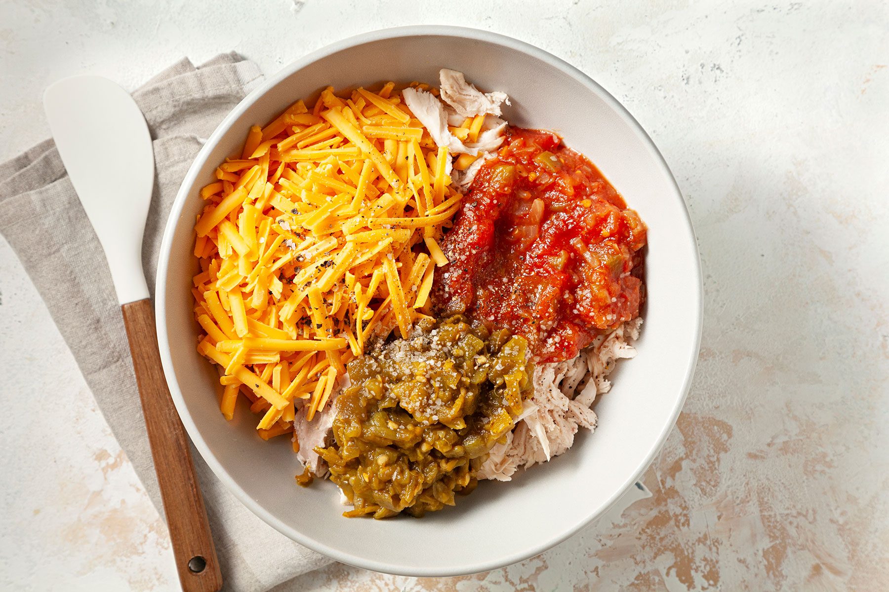 A white bowl on a beige napkin contains shredded chicken, grated cheddar cheese, red salsa, and green chilies. A wooden spoon rests nearby.