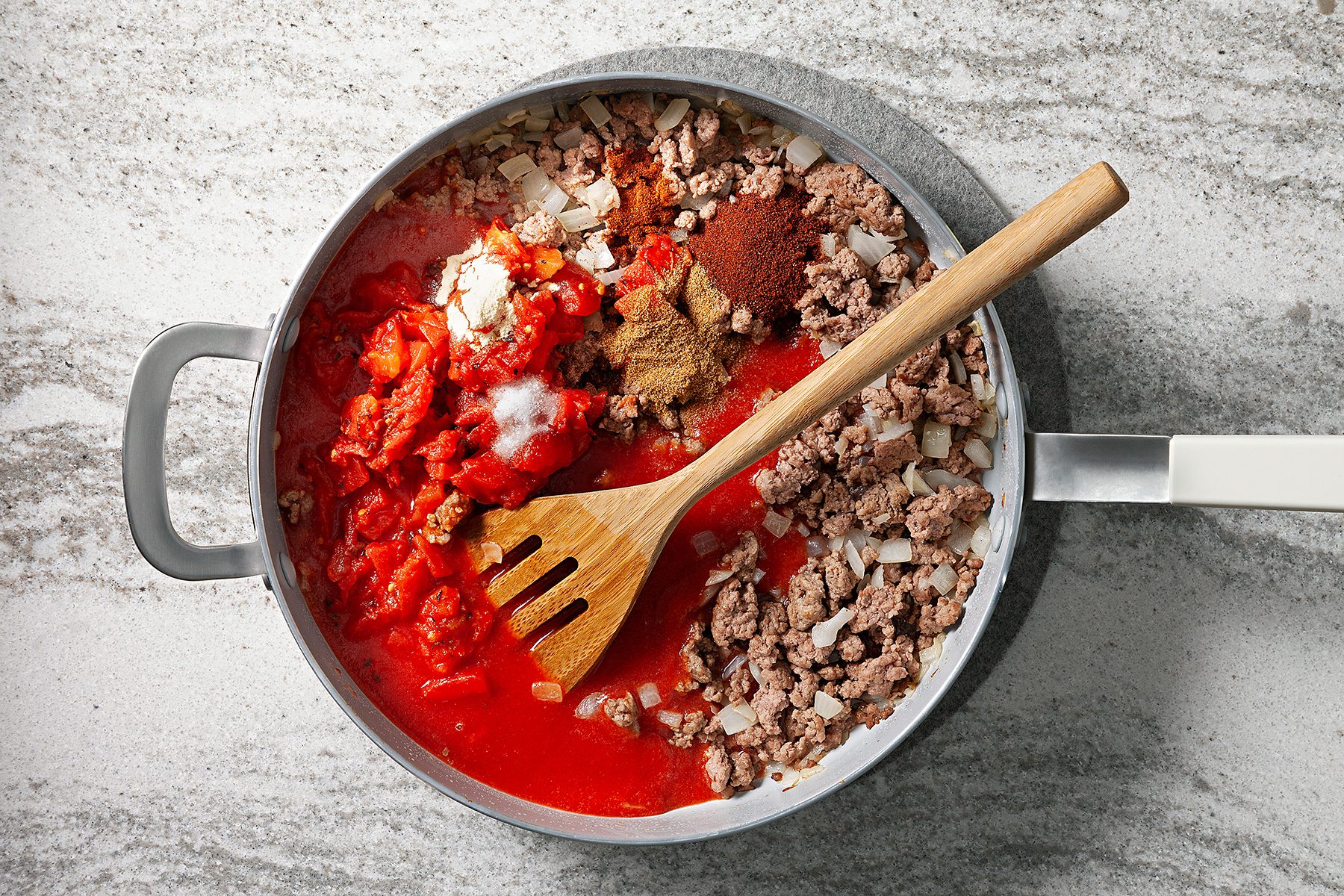 A frying pan on a marble surface contains ingredients for a meal, including ground meat, chopped tomatoes, onions, and various spices. A wooden spatula rests inside the pan.