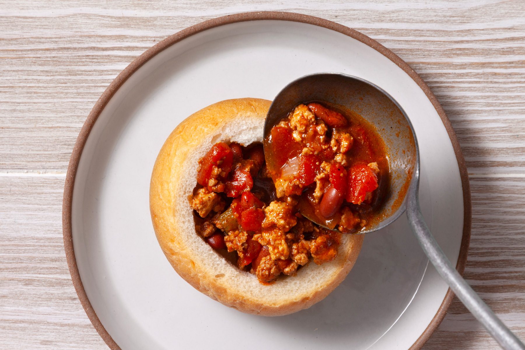 An overhead shot of a bowl of chili being poured into a hollowed-out bread roll. The chili features a thick, red sauce with visible chunks of meat and vegetables. The bread roll is golden brown and has a soft texture.