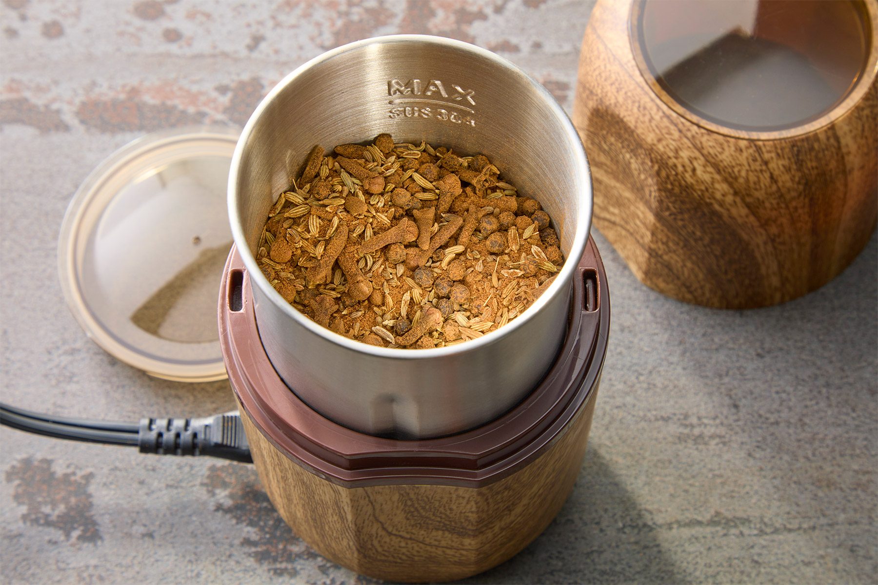 A wooden coffee grinder with an open lid contains freshly ground coffee beans. A power cord is attached, and an additional wooden lid is visible nearby on a gray surface.