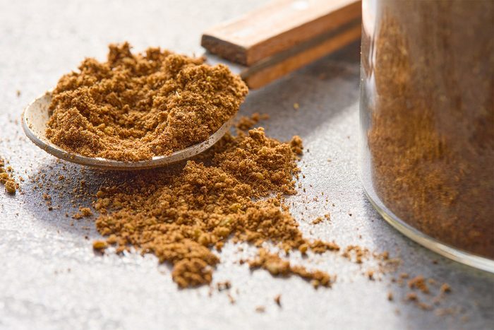 A close-up of a spoon filled with ground cinnamon, with some of the spice spilled on a textured surface. To the right, part of a glass jar containing more cinnamon is visible. The scene is lit with soft, natural light.