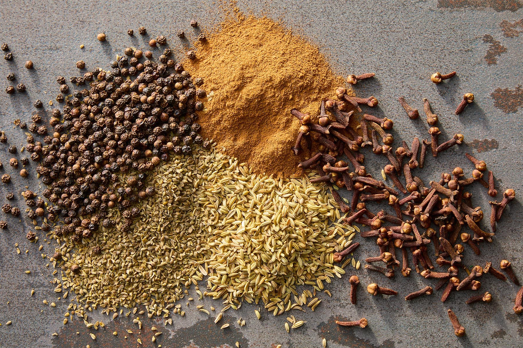 A flat lay of assorted spices on a gray surface, featuring a pile of black peppercorns, ground cinnamon, fennel seeds, and whole cloves, arranged in a visually appealing manner.