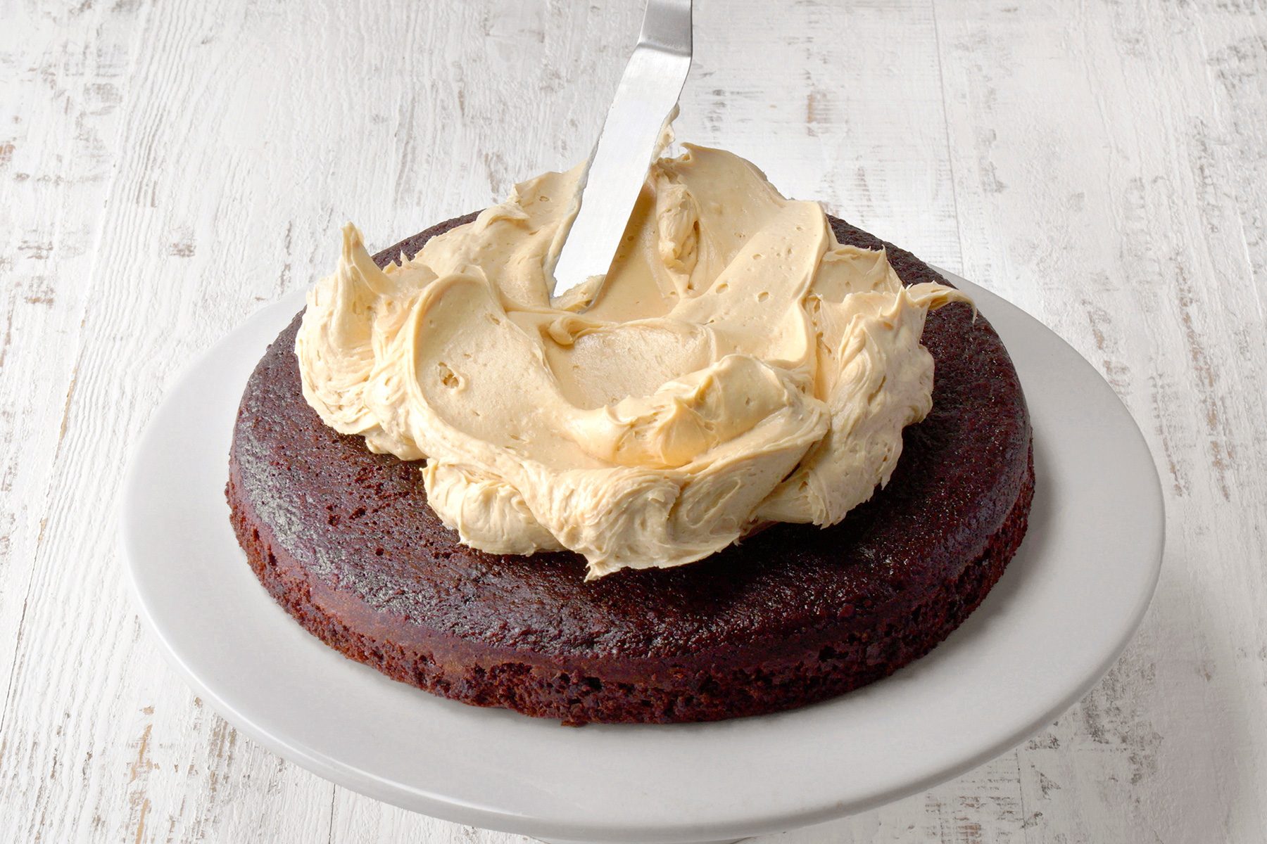 A chocolate cake on a white plate is being frosted with light brown icing. A spatula is smoothing the icing on top of the cake, which sits on a white wooden surface.
