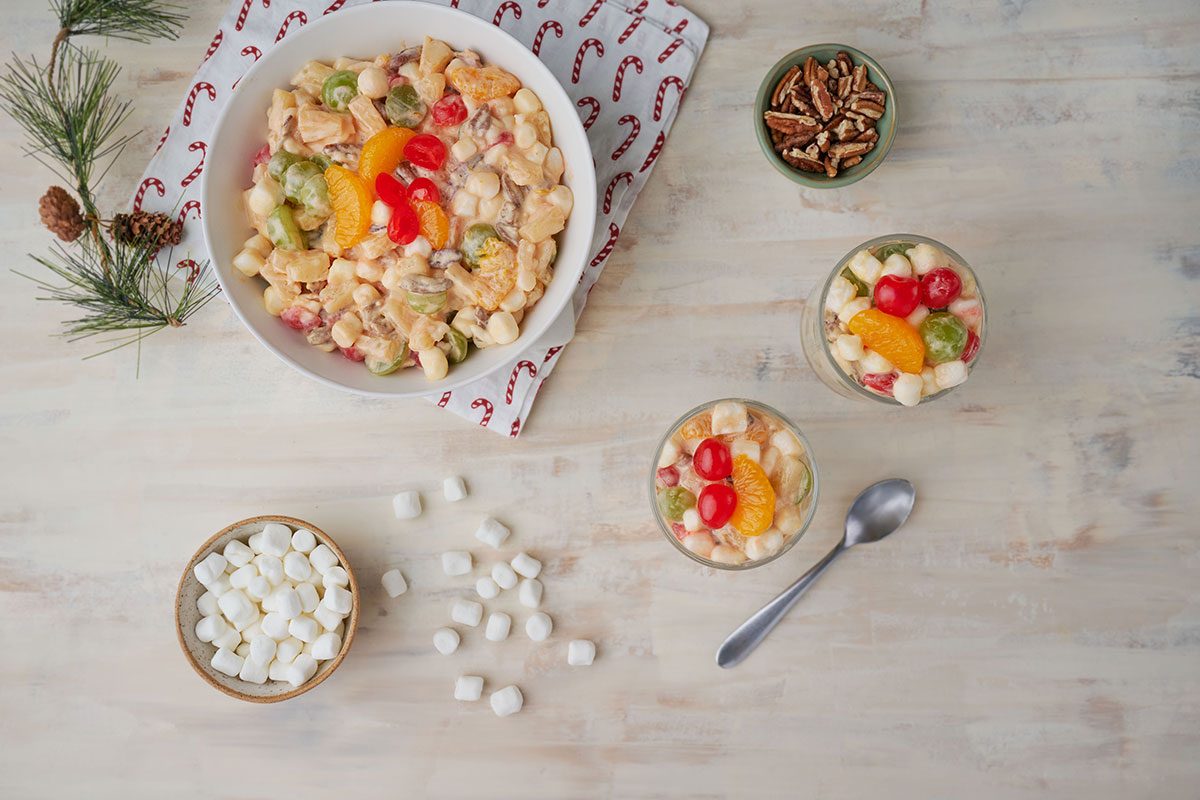 Overhead shot of a serving of Christmas Fruit Salad in a glass bowl, accompanied by marshmallows and pecans on the side for additional garnish.