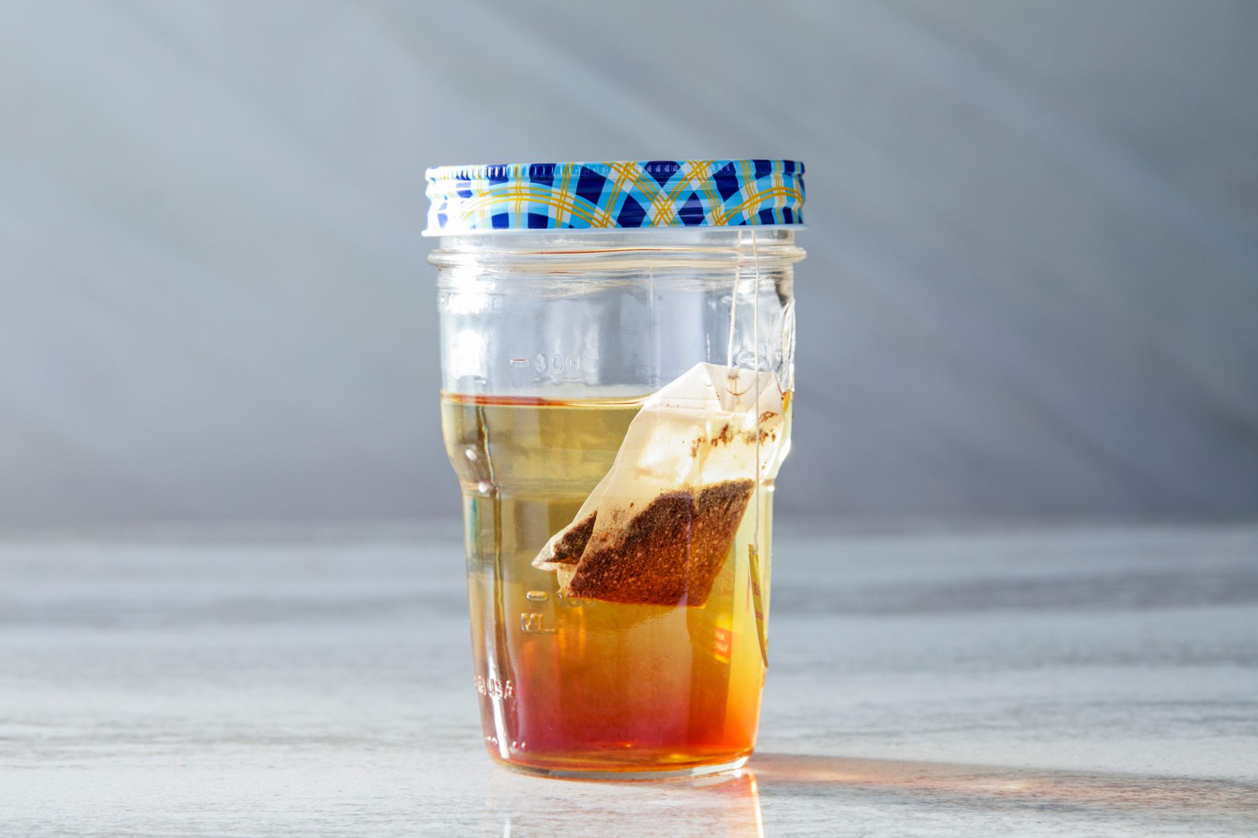 Horizontal shot of a glass jar with a blue and yellow lid filled with tea, featuring a mostly submerged tea bag inside. The jar is placed on a light grey countertop.