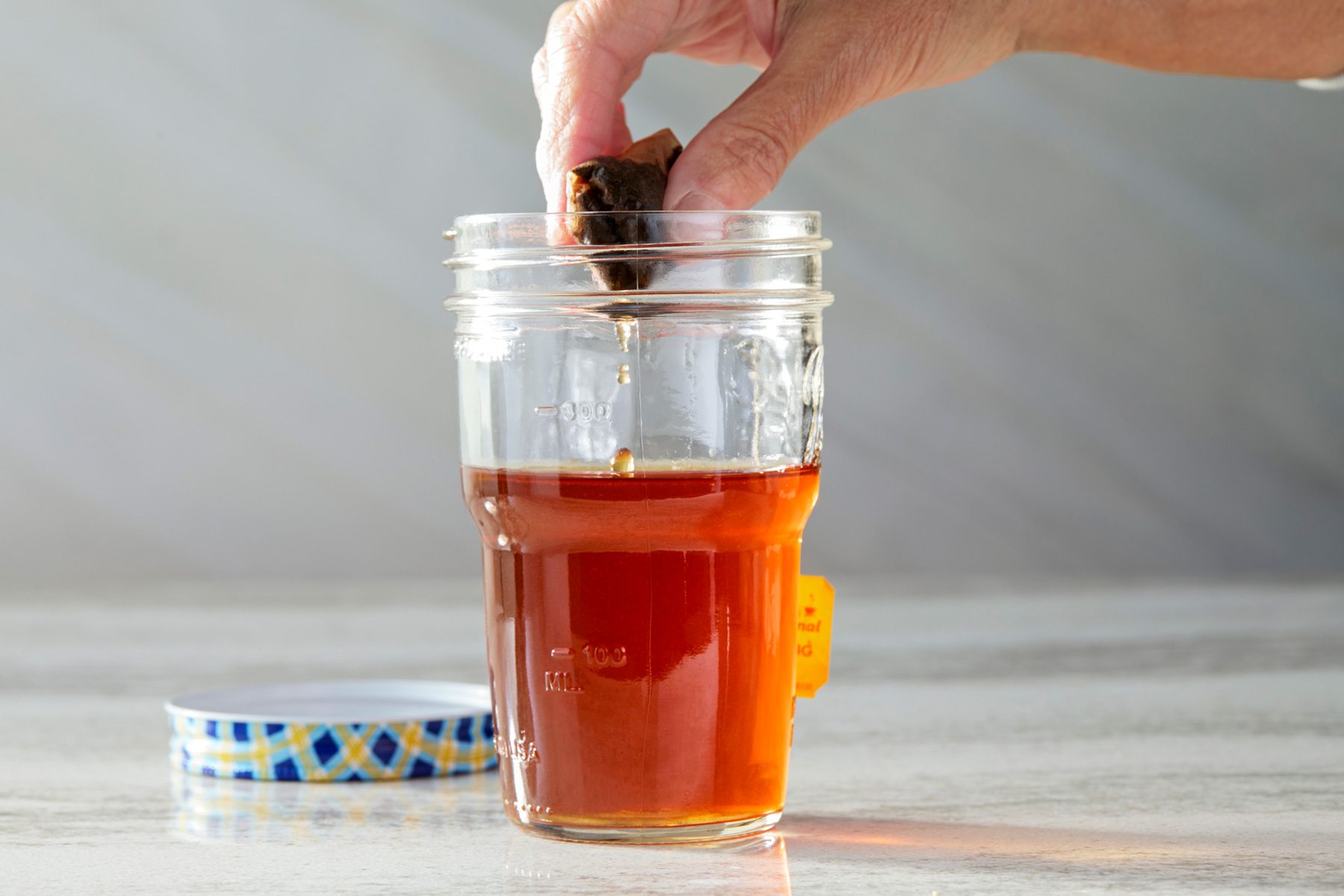 Horizontal shot of a hand dropping a tea bag into a glass jar with brewed tea. The jar, featuring a lid with a blue, yellow, and white design, contains deep amber tea with a small amount left. The tea bag is partially submerged, and the background is light gray.