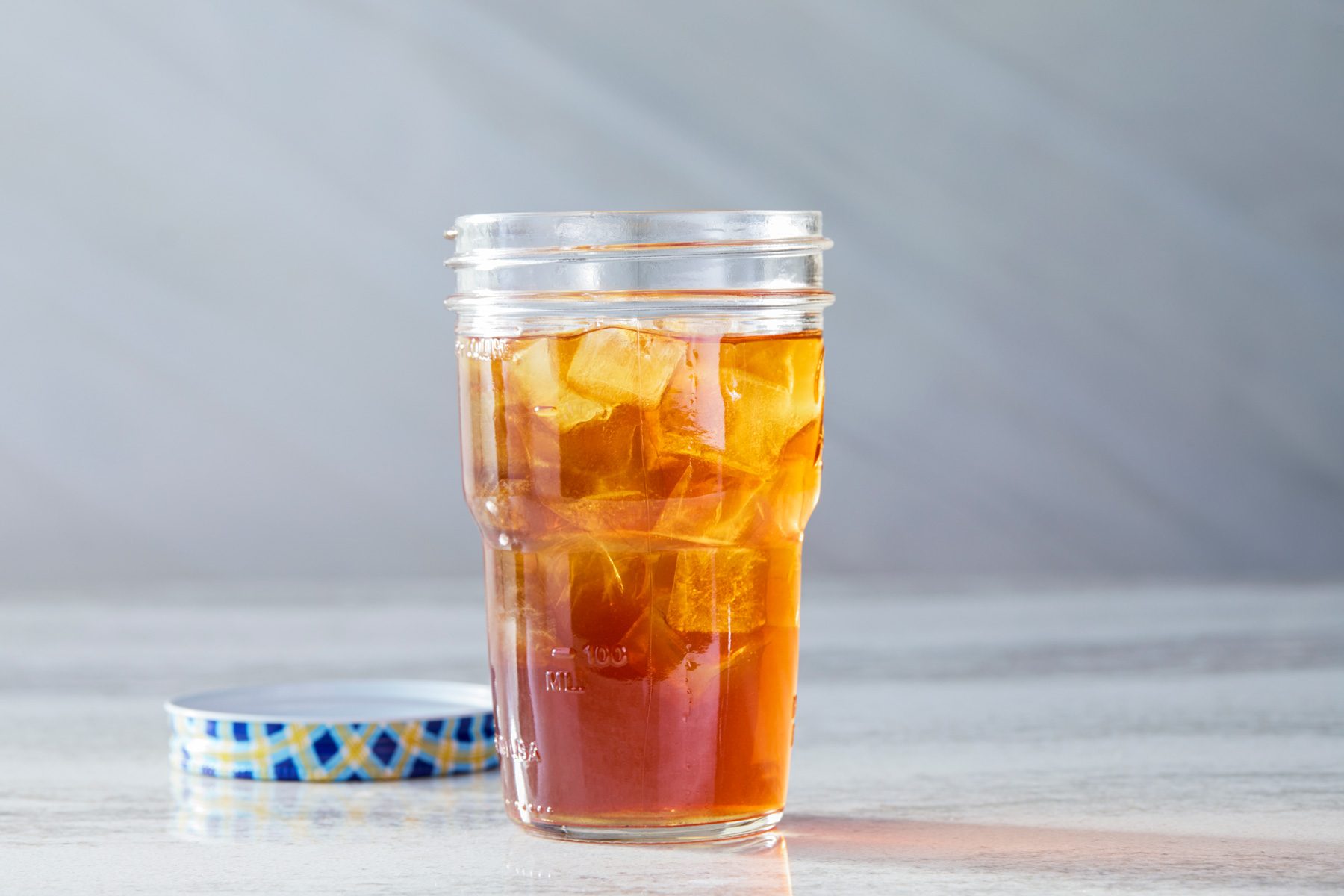 Horizontal shot of a clear glass jar filled with amber-colored iced tea on a light gray surface featuring a veined pattern. The jar has a lid with a blue and gold design, and ice cubes are floating in the tea.