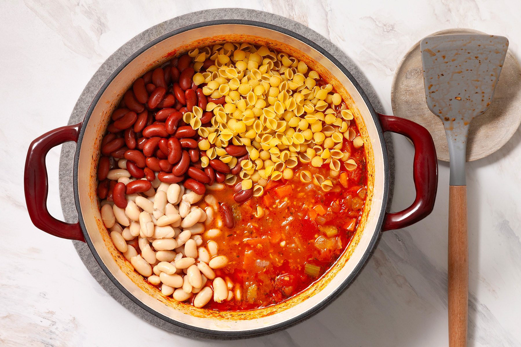 A pot overflows with uncooked pasta, red kidney beans, white beans, and a tomato-based vegetable stew. A spatula rests on a stone dish nearby, on a white countertop.