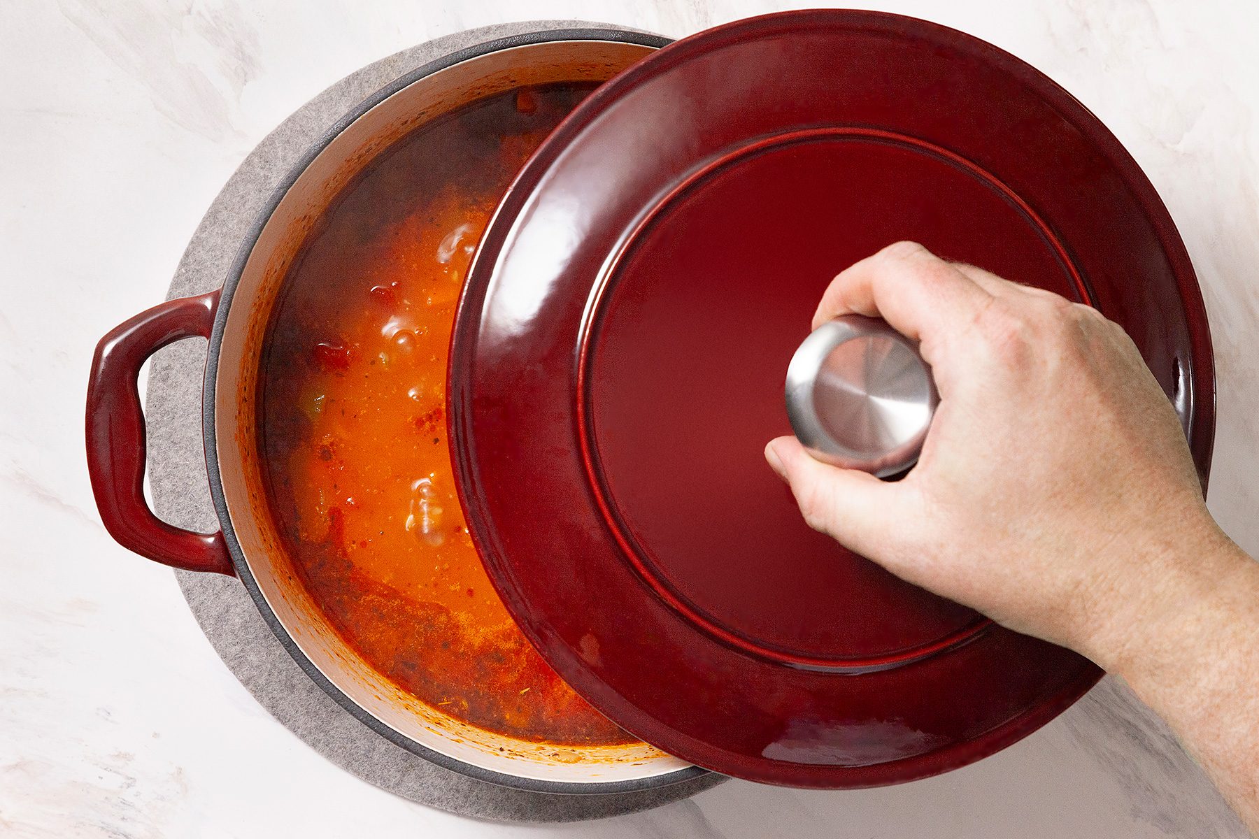 A hand lifts the lid of a red pot, revealing a simmering stew with visible pieces of meat and vegetables. The pot rests on a light-colored surface.