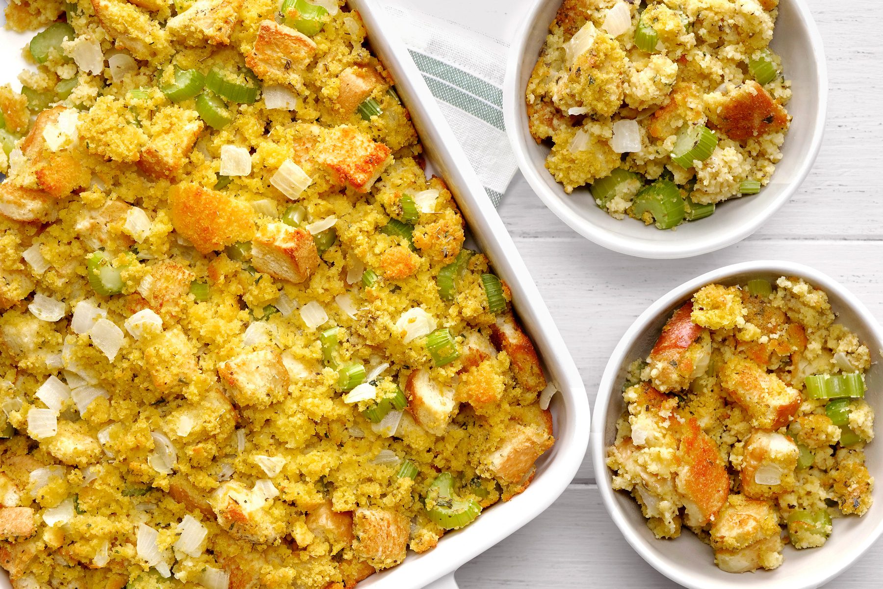 overhead shot of a white baking dish filled with Cornbread Dressing is showcased, surrounded by two smaller bowls containing individual servings. The stuffing is packed with texture and flavor, featuring cubes of cornbread, chopped celery, and grated Parmesan cheese;