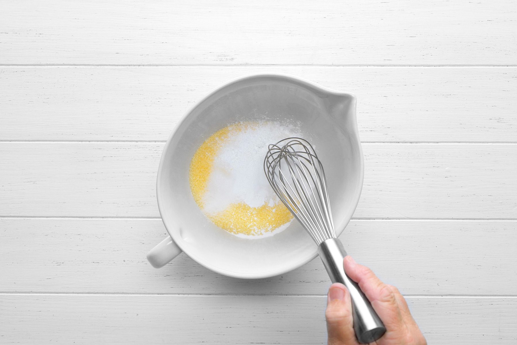overhead shot of A white bowl contains a mixture of melted butter and a white powder, likely flour, A whisk is being used to stir the mixture together, The mixture is a pale yellow color and is starting to bubble slightly, The bowl is sitting on a wooden surface;