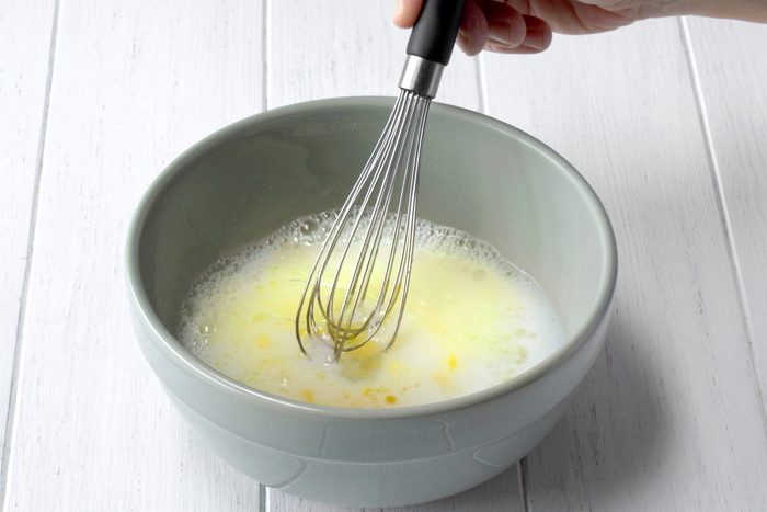 3/4th shot of a hand holding a whisk and whisking a mixture in a gray bowl, The mixture appears to be a light yellow color and has bubbles forming on the surface, The bowl is placed on a white wooden surface;