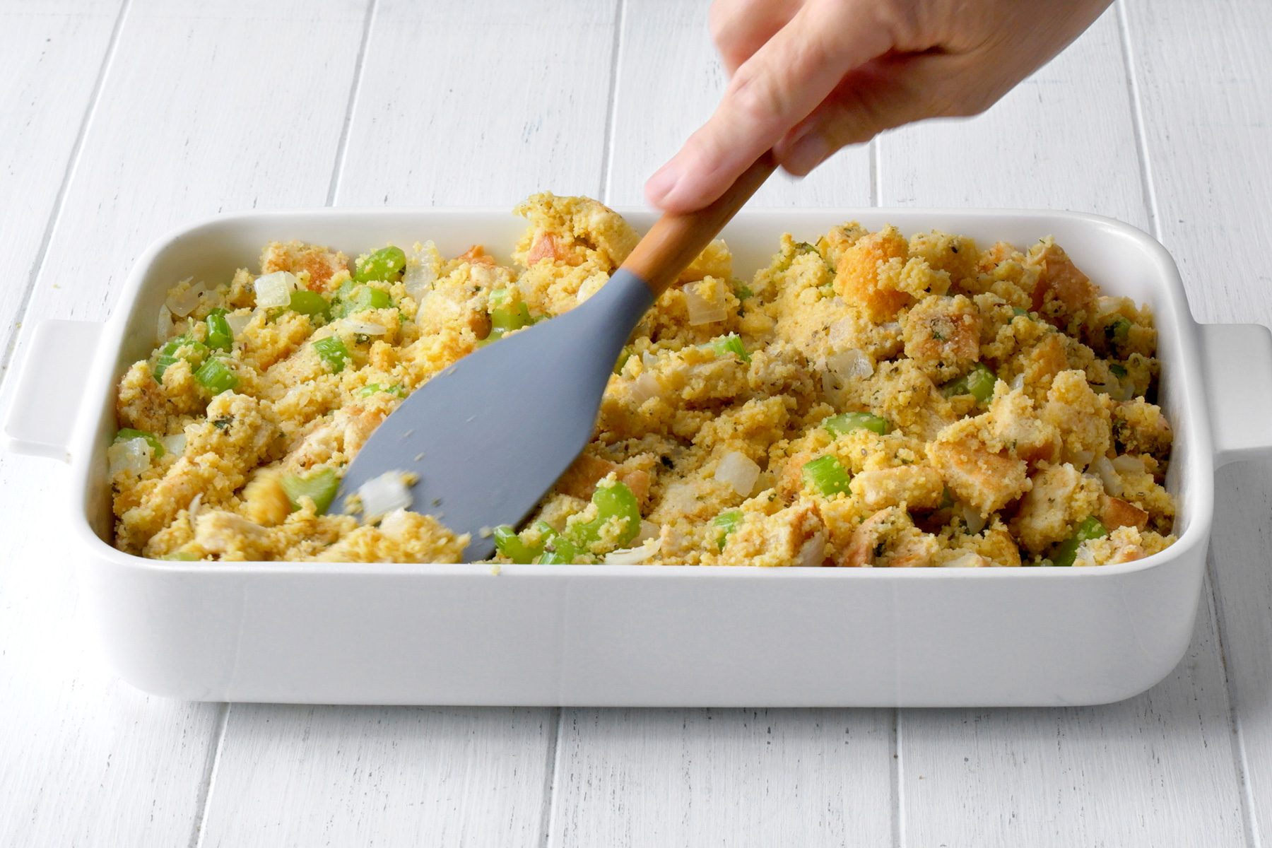 3/4th shot of a person is using a spatula to spread a mixture of cornbread, vegetables, and spices in a white baking dish, The mixture is a light brown color and contains visible pieces of cornbread, chopped celery, and onion, The baking dish is sitting on a white wooden surface;