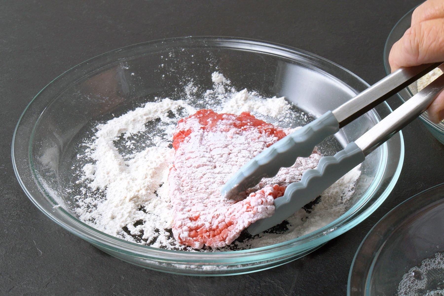 3/4th shot of a hand using tongs to coat a piece of meat in flour, The meat is a reddish-brown color and is partially covered in a white layer of flour, The meat is resting in a glass bowl filled with flour, The bowl is placed on a dark gray surface