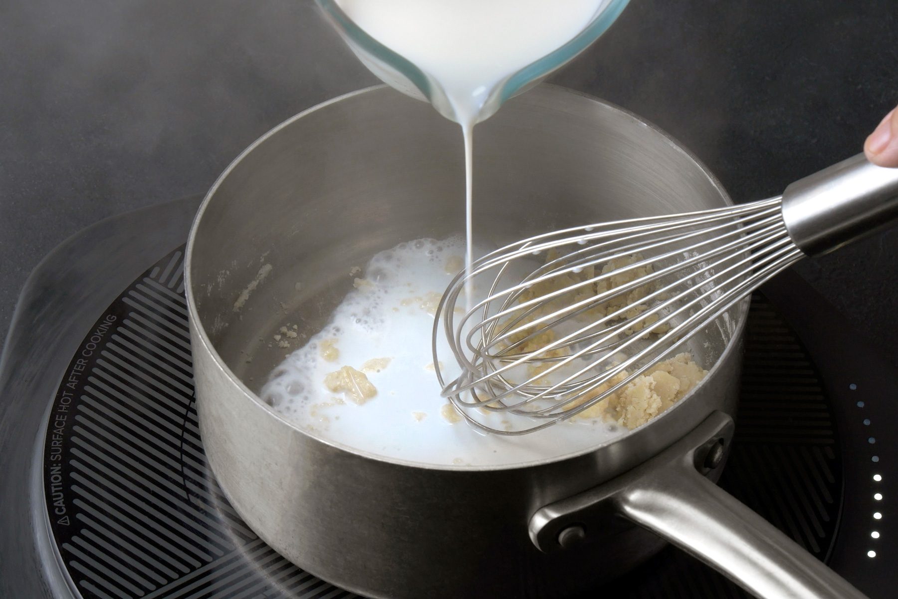 3/4th shot of a hand pouring a liquid into a saucepan on an electric induction cooktop. The saucepan contains a mixture of melted butter and flour, and the liquid is being added gradually while whisking; The liquid appears to be milk or cream, and the mixture is starting to thicken;