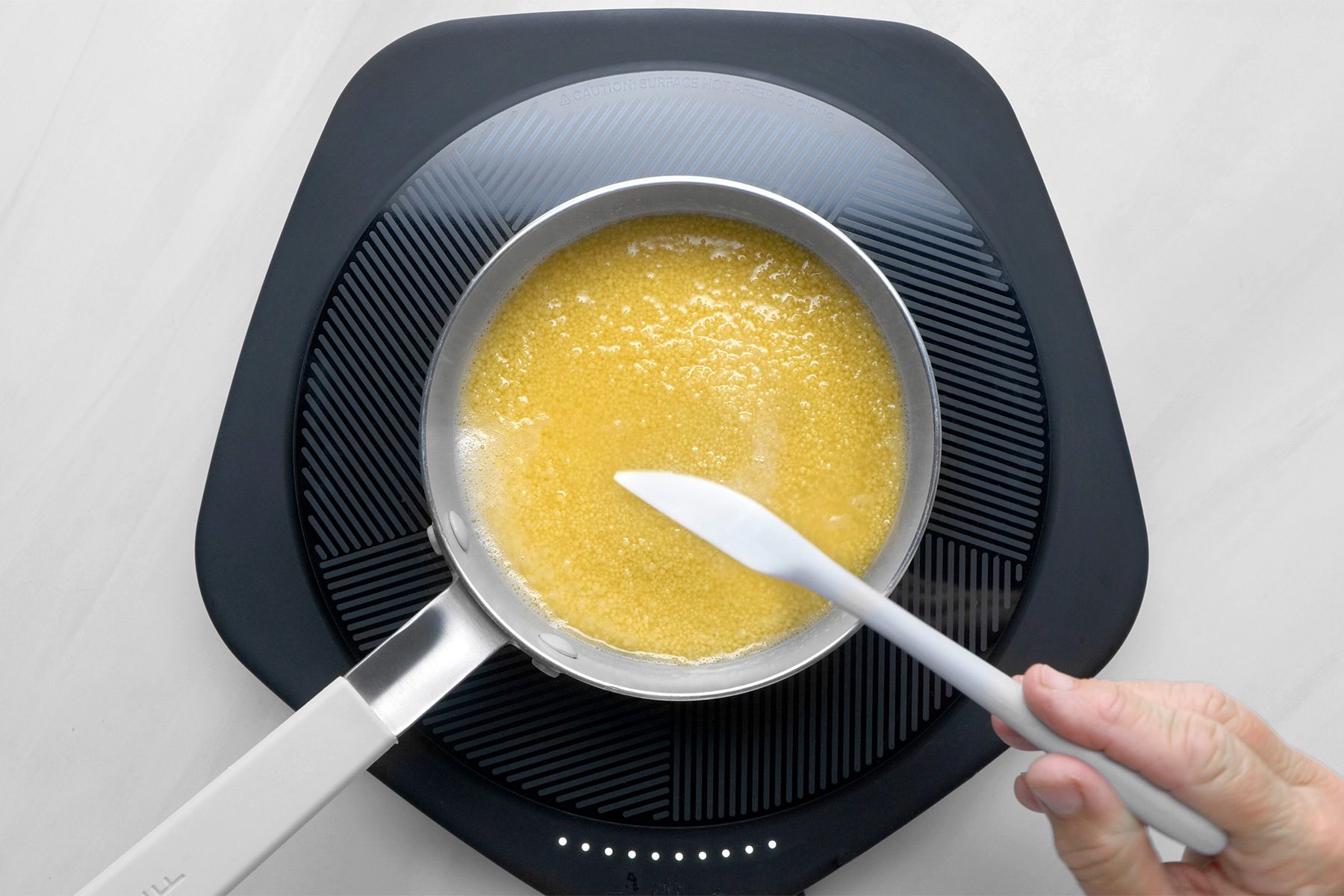 overhead shot of a small saucepan on an electric induction cooktop, The saucepan contains melted butter, which is a pale yellow color and has a smooth texture, The butter is bubbling slightly, indicating that it is heating up, A spatula is being used to stir the butter, ensuring it is evenly melted;