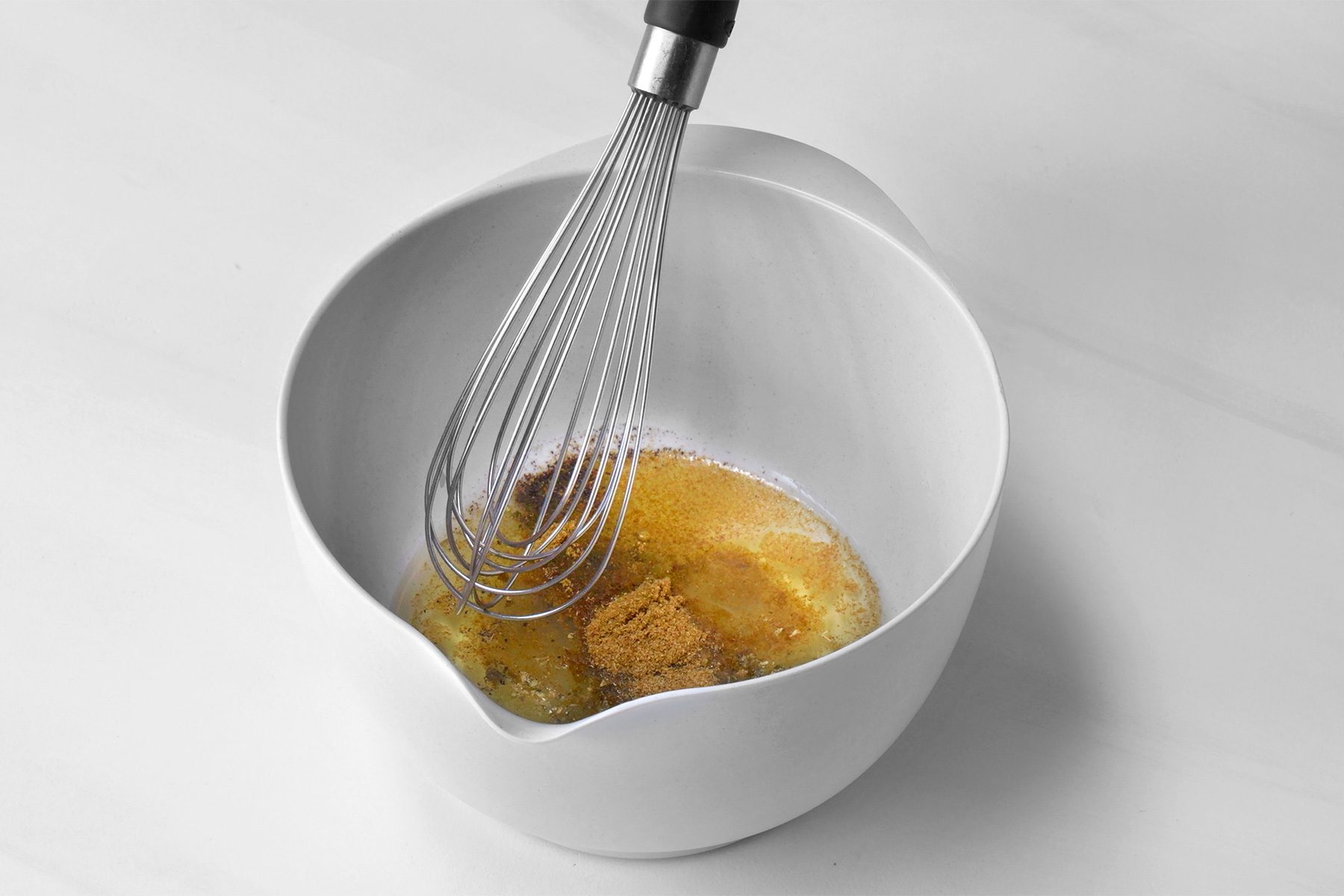 3/4th shot of a white bowl contains a mixture of melted butter, brown sugar, and spices, A whisk is being used to stir the ingredients together, creating a creamy and fragrant sauce;