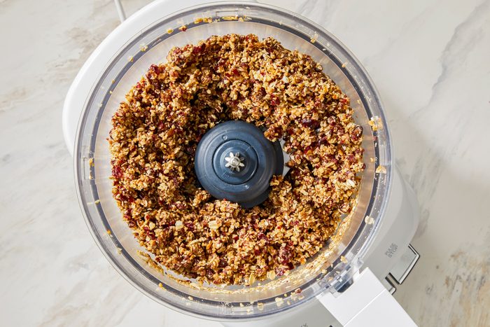 A food processor with the lid on, showing a mixture of oats, nuts, and dried fruits being pulsed.