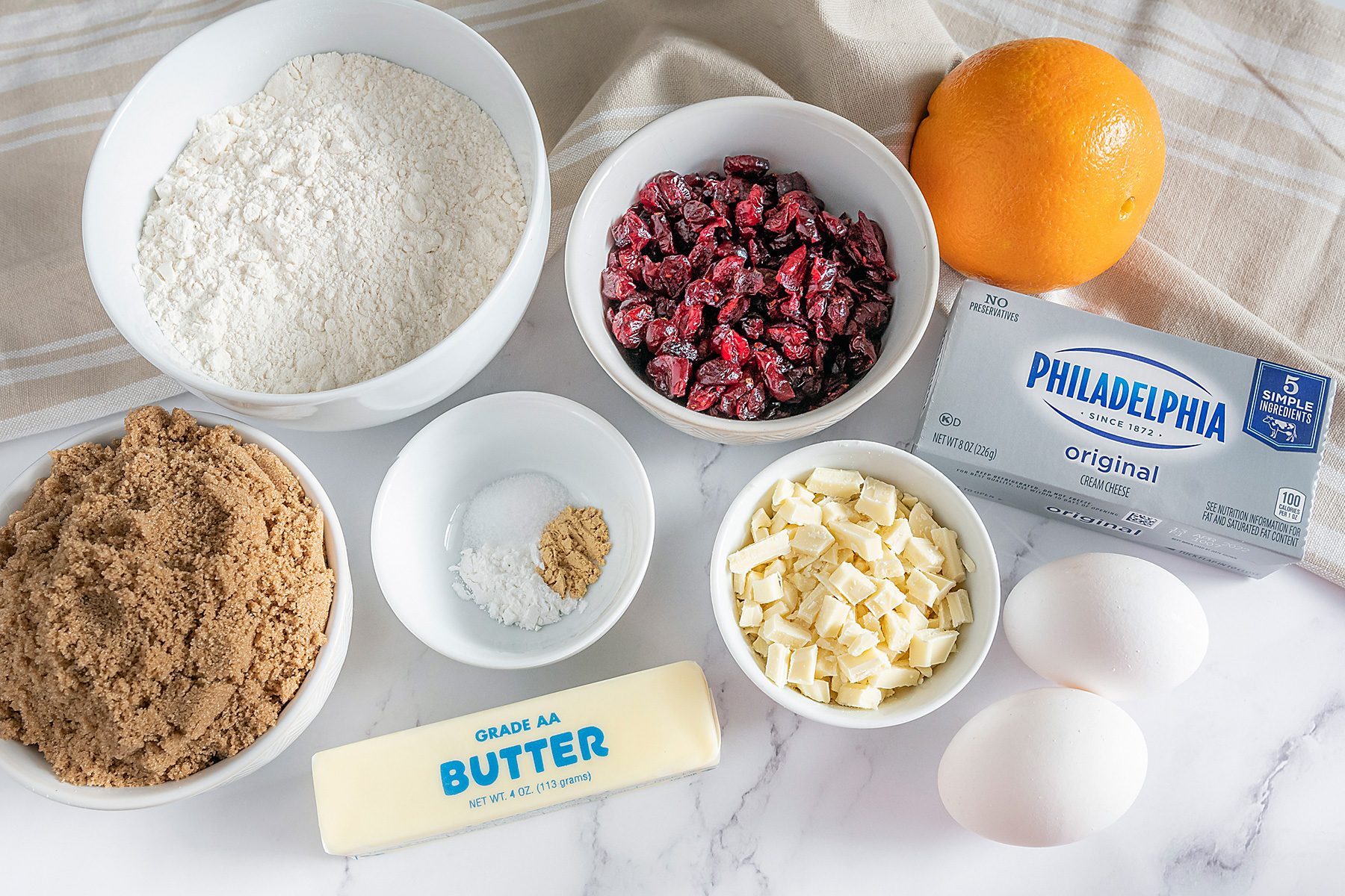 Baking ingredients on a marble surface: flour, dried cranberries, an orange, brown sugar, baking soda, eggs, white chocolate chips, a stick of butter, and a block of cream cheese.